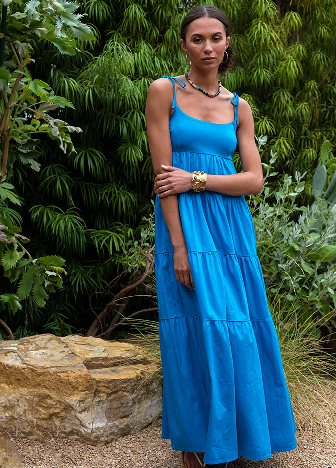 Model wears blue maxi dress with tie straps standing in front of greenery.