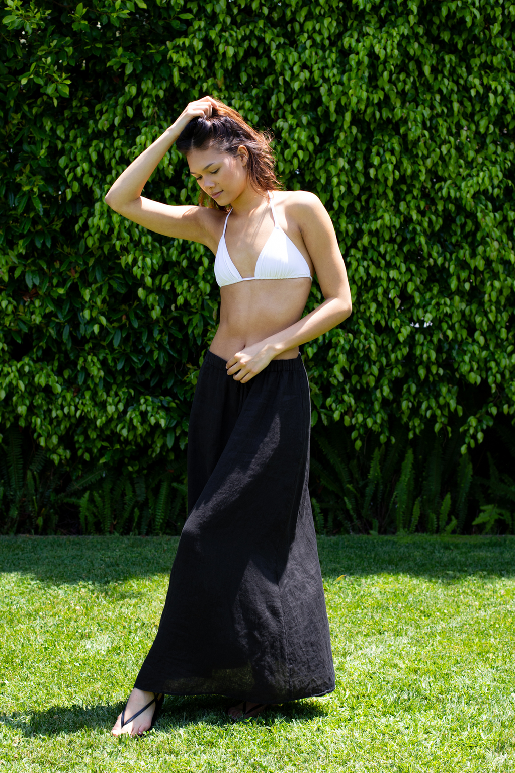 MODEL STANDING ON GRASS WITH GREENERY BEHIND HER. HER LEGS ARE CROSSED AND SHE IS WEARING A WHITE BIKINI TOP AND BLACK MAXI DRAWSTRING LINEN SKIRT. 