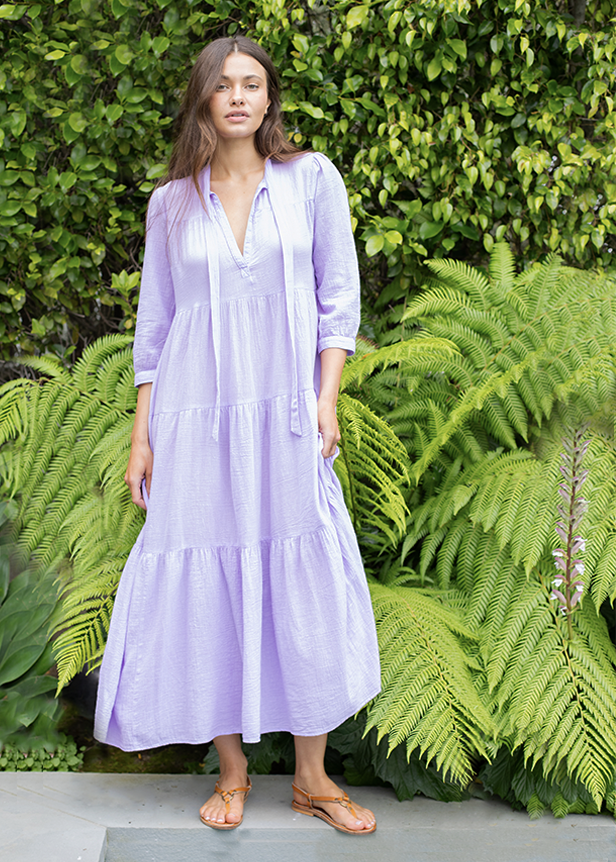 Model wears long lavendar v neck dress with long sleeves and tan sandals. She stands outside in front of greenery.